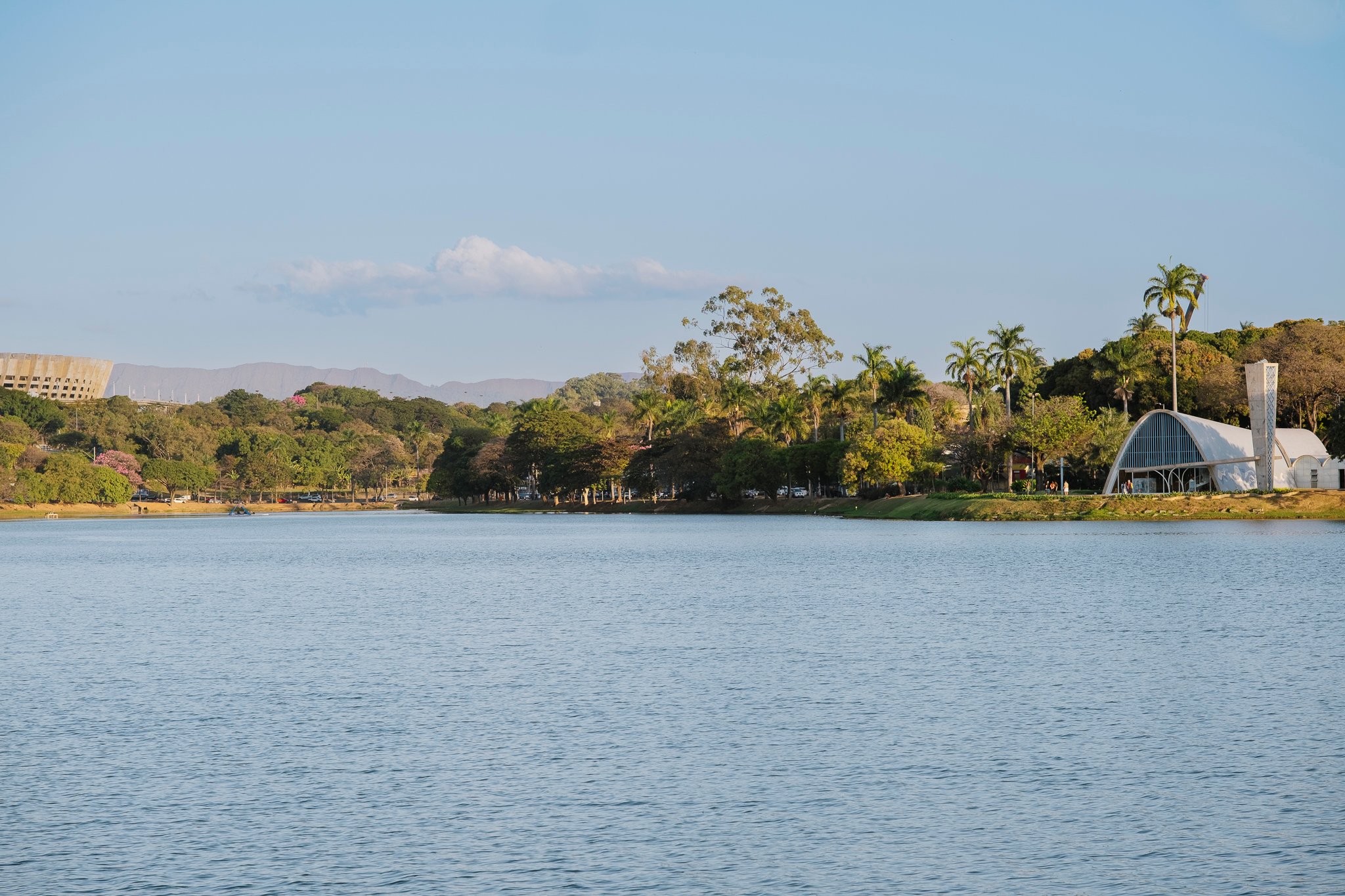 Calm Pampulha Lagoon with modernist São Francisco de Assis Church and palm trees at dusk, Mineirão stadium in distance.