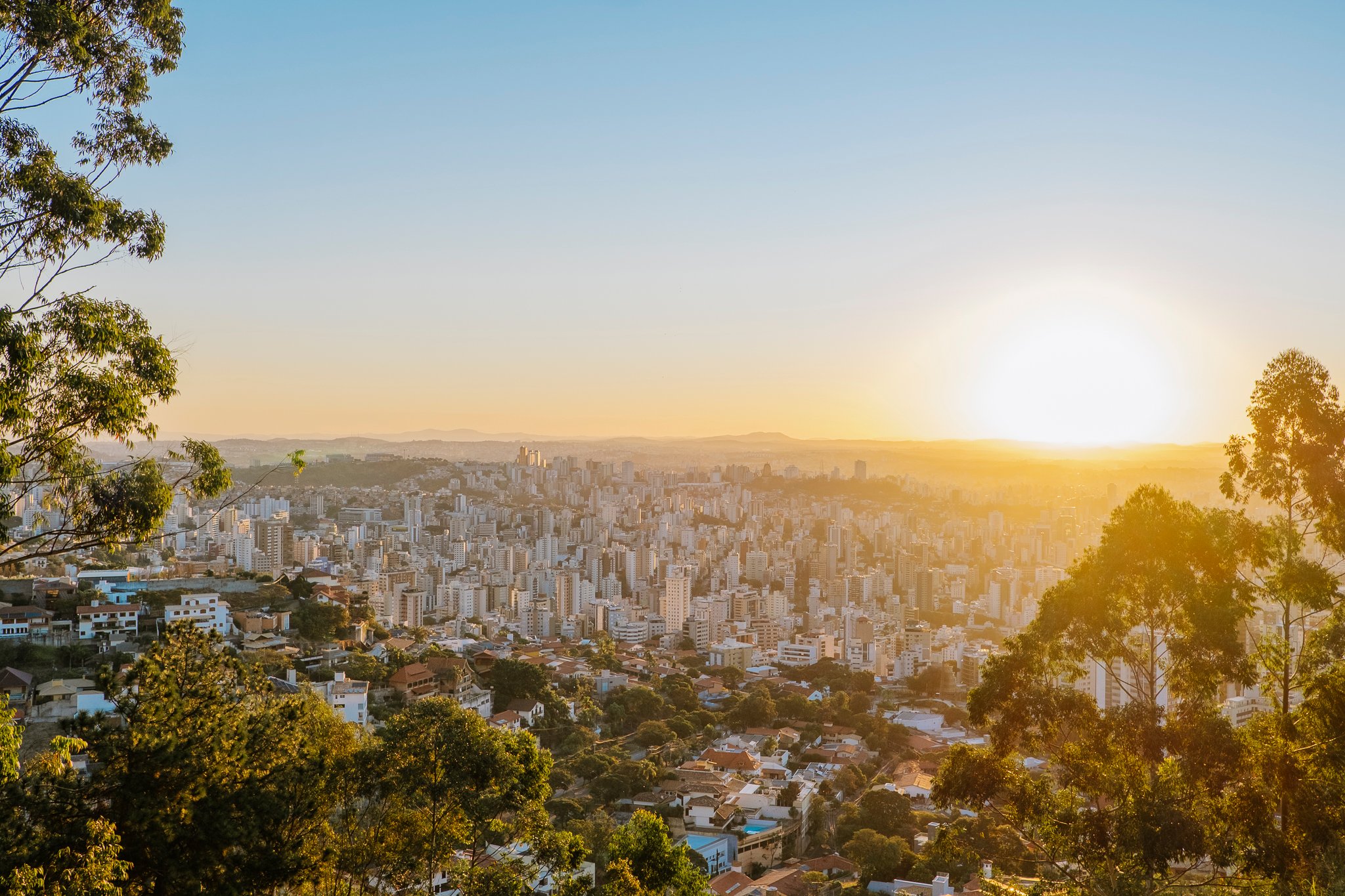 Wide sunset view over Belo Horizonte skyline seen from Mirante do Mangabeiras, framed by eucalyptus trees.
